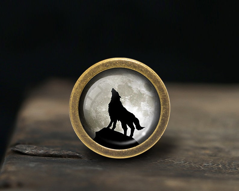 Może przedstawiać: Okrągły, mosiężny, ozdobny gałka z wkładką szklaną przedstawiającą czarną sylwetkę wilka wyjącego do pełnego księżyca.
