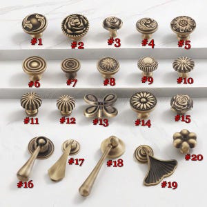 Pu&ograve; includere: Una collezione di pomelli e maniglie per armadi in ottone antico. I disegni includono forme floreali, geometriche e a farfalla. Ogni pomello &egrave; numerato da #1 a #20. L'hardware &egrave; esposto su una superficie bianca.