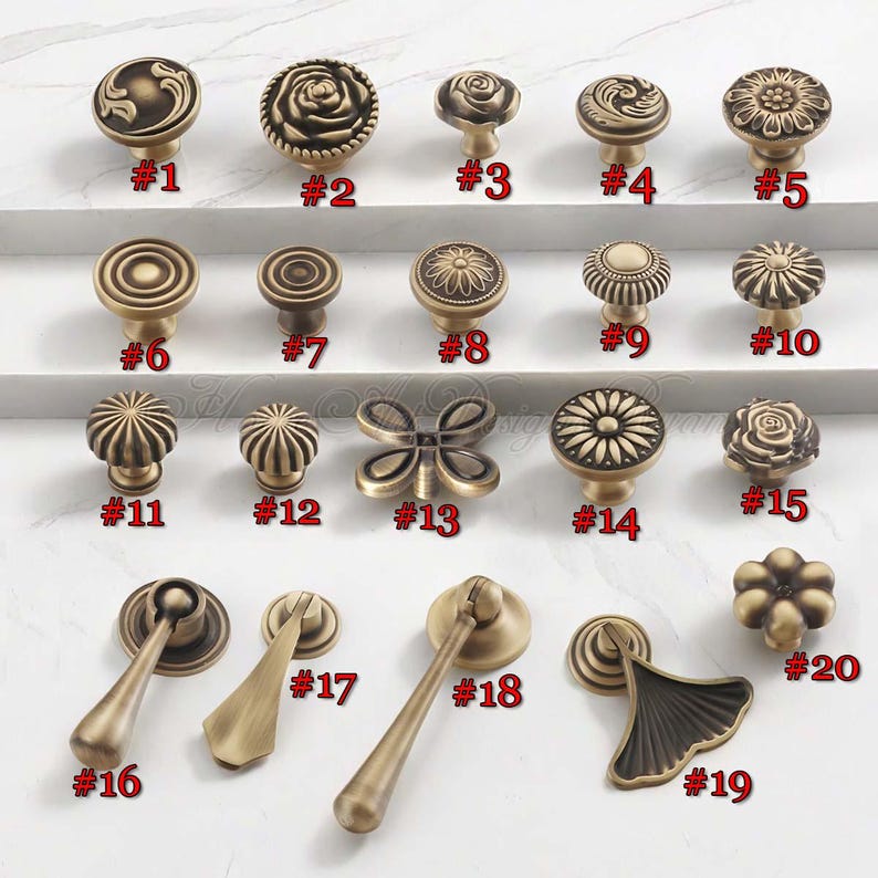 Pu&ograve; includere: Una collezione di pomelli e maniglie per armadi in ottone antico. I disegni includono forme floreali, geometriche e a farfalla. Ogni pomello &egrave; numerato da #1 a #20. L'hardware &egrave; esposto su una superficie bianca.