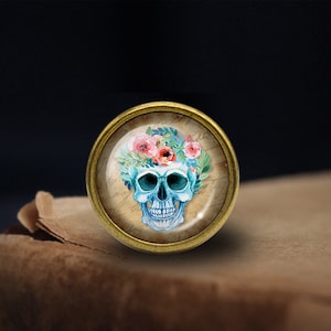 May include: A round, gold-toned metal button with a glass top featuring a blue and white skull with a floral crown of pink and white flowers. The skull has a detailed design with a floral pattern on the top of the head.