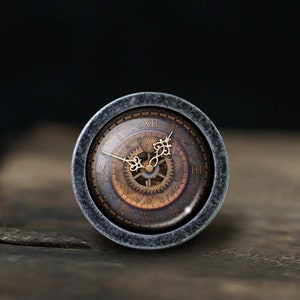 Puede incluir: Una esfera de reloj redonda de color gris con números romanos y engranajes intrincados. La esfera está rodeada por un marco de metal gris.