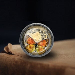 Pode incluir: Um bot&atilde;o de prata com uma parte superior de vidro que apresenta uma ilustra&ccedil;&atilde;o vintage de uma borboleta laranja sobre um fundo creme com acentos pretos ornamentados.