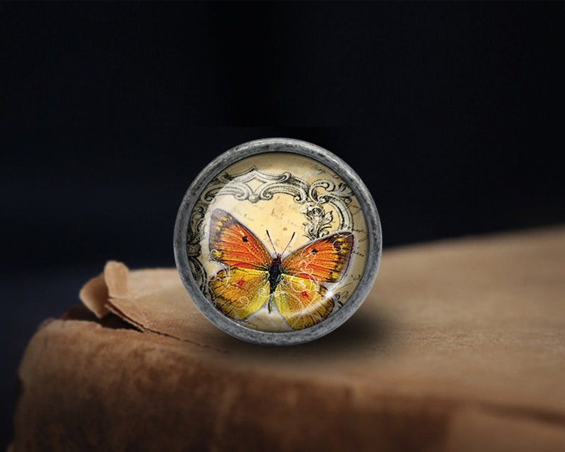Pode incluir: Um bot&atilde;o de prata com uma parte superior de vidro que apresenta uma ilustra&ccedil;&atilde;o vintage de uma borboleta laranja sobre um fundo creme com acentos pretos ornamentados.