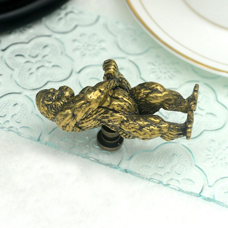 Puede incluir: Un pomo de armario de metal dorado con forma de gorila. El gorila est&aacute; sentado en cuclillas con los brazos levantados por encima de la cabeza.