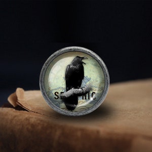 Op de afbeelding: Een ronde zilverkleurige knop met een glazen inzet met daarop een zwarte raaf die op een tak zit. De glazen inzet heeft een blauwe en witte achtergrond met het woord "SEPHIC" erop gedrukt.