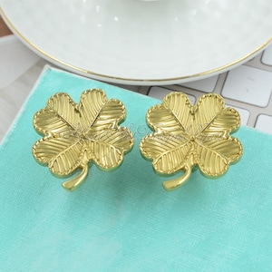 Puede incluir: Dos pomos decorativos dorados en forma de trébol de cuatro hojas. Los pomos tienen patrones detallados de venas de hojas y un tallo curvo. Se colocan sobre un fondo azul claro, con un plato blanco en el fondo.