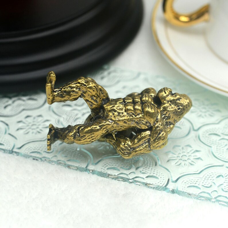 K&ouml;nnte beinhalten: Eine goldfarbene Metallfigur eines kleinen, hockenden Wesens mit einer strukturierten Oberfl&auml;che. Die Figur steht auf einer Glasfl&auml;che mit einem Blumenmuster.