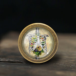 Könnte beinhalten: Ein runder Knopf in antikem Messing-Finish, der eine detaillierte Abbildung eines menschlichen Brustkorbs zeigt, der mit bunten Blumen und Blättern gefüllt ist. Der Knopf hat eine klare, leicht gewölbte Oberfläche.