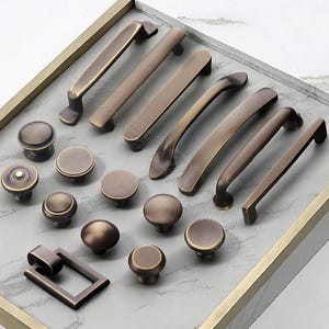 K&ouml;nnte beinhalten: Eine Auswahl an bronzefarbener Schrankbeschl&auml;ge, darunter verschiedene Griffe und Kn&ouml;pfe. Die Griffe variieren in der L&auml;nge, und die Kn&ouml;pfe sind rund. Die Beschl&auml;ge sind auf einer hellgrauen Oberfl&auml;che in einem goldfarbenen Tablett angeordnet.