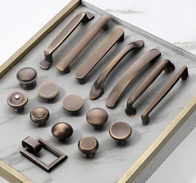 K&ouml;nnte beinhalten: Eine Auswahl an bronzefarbener Schrankbeschl&auml;ge, darunter verschiedene Griffe und Kn&ouml;pfe. Die Griffe variieren in der L&auml;nge, und die Kn&ouml;pfe sind rund. Die Beschl&auml;ge sind auf einer hellgrauen Oberfl&auml;che in einem goldfarbenen Tablett angeordnet.