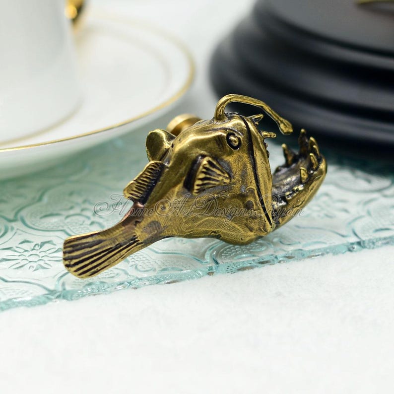 K&ouml;nnte beinhalten: Detaillierte Messingfarbene Anglerfisch-Figur. Die Skulptur zeigt ein offenes Maul mit sichtbaren Z&auml;hnen, Flossen und einem K&ouml;der. Der Fisch befindet sich auf einer transparenten, strukturierten Oberfl&auml;che, mit einer wei&szlig;en Tasse und Untertasse im Hintergrund.