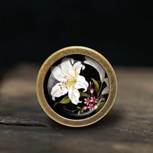 K&ouml;nnte beinhalten: Ein runder, messingfarbener Knopf mit einer wei&szlig;en Lilie und rosa Bl&uuml;ten auf schwarzem Hintergrund.