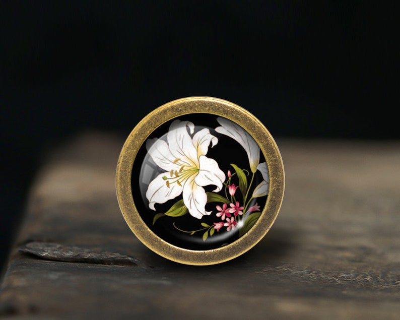 K&ouml;nnte beinhalten: Ein runder, messingfarbener Knopf mit einer wei&szlig;en Lilie und rosa Bl&uuml;ten auf schwarzem Hintergrund.