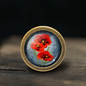 May include: A round, brass-coloured knob with a glass insert featuring a floral design of three red poppies on a blue background.