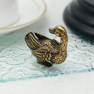 Op de afbeelding: Een klein, goudkleurig metalen beeldje van een eend met zijn kop naar achteren gedraaid. De eend staat op een helder glazen oppervlak met een decoratief patroon.