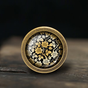 May include: A round, brass-colored button with a black and gold floral design. The button has a raised edge and a smooth, shiny surface.