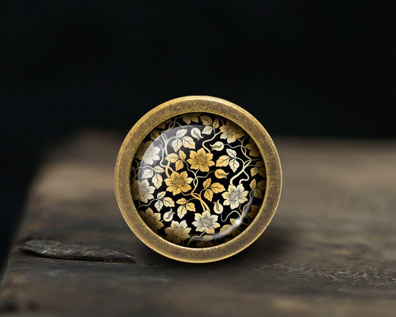 May include: A round, brass-colored button with a black and gold floral design. The button has a raised edge and a smooth, shiny surface.