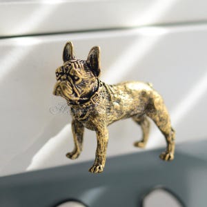 Puede incluir: Tirador de cajón de metal dorado en forma de bulldog francés. El bulldog está de pie sobre sus cuatro patas con la cabeza ligeramente inclinada hacia un lado.