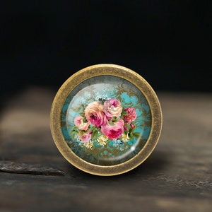 以下が含まれることがあります： アンティークな花柄が施されたガラス製のインサートがついた、真鍮色の丸いノブ。デザインには、ピンクと白のバラ、青い勿忘草、白いデイジーが、ターコイズブルーの背景に金色の模様で描かれています。