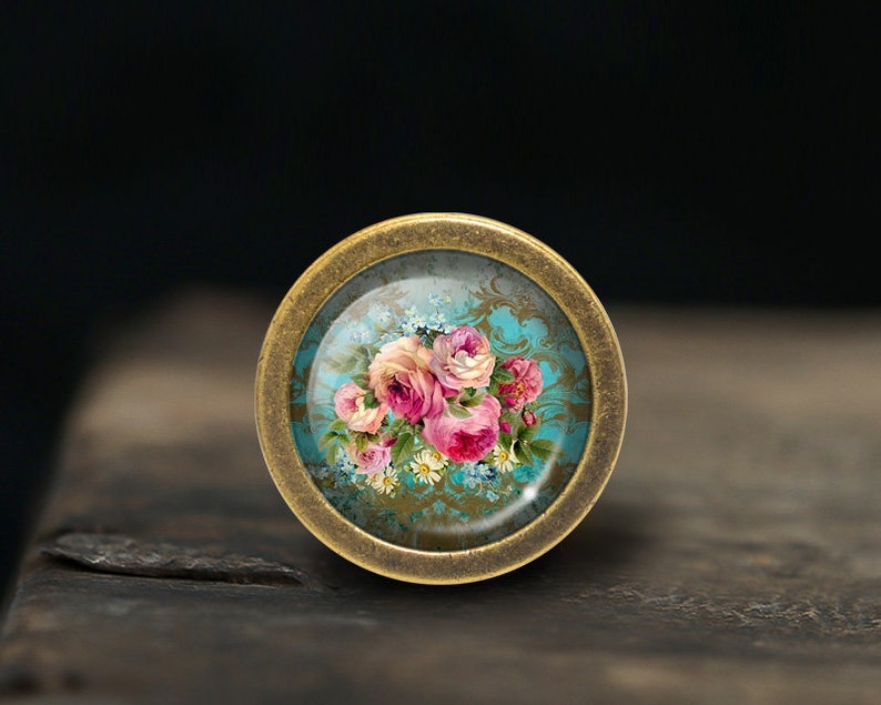 May include: A round, brass-colored knob with a glass insert featuring a vintage floral design. The design includes pink and white roses, blue forget-me-nots, and white daisies on a teal background with a gold pattern.