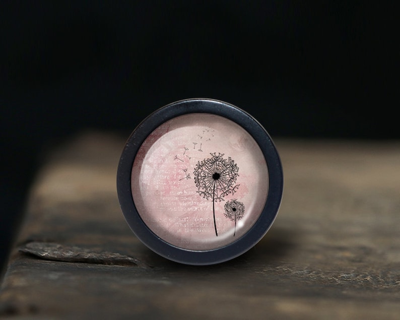 Peut inclure: Un pendentif rond noir avec un fond rose et blanc repr&eacute;sentant une silhouette noire de deux pissenlits avec des graines qui s'envolent dans le vent.