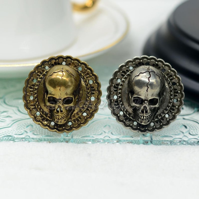 May include: Two decorative skull-shaped embellishments. One is gold-toned, the other silver-toned, each set within a detailed, circular frame. The skulls have visible eye sockets and teeth, with a cracked appearance on the silver skull.