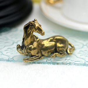 Könnte beinhalten: Eine kleine, goldfarbene, bronzene Figur eines liegenden Pferdes. Das Pferd befindet sich in einer entspannten Pose mit dem Kopf zur Seite gedreht.