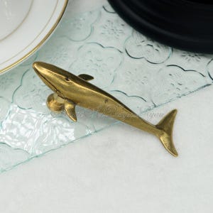 May include: A gold-colored whale figurine, with a detailed design, is displayed on a clear glass surface with a decorative pattern.