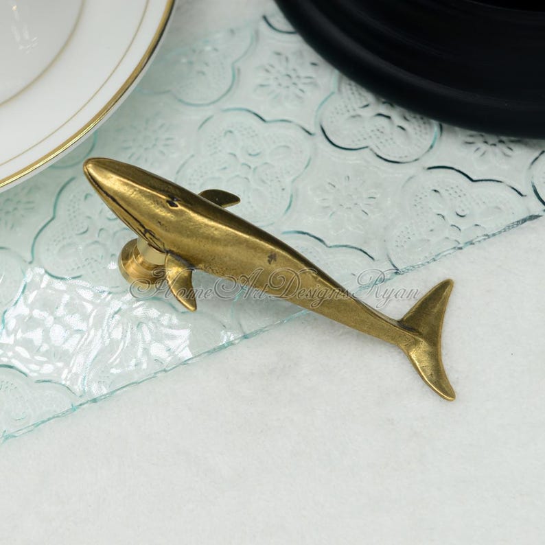 May include: A gold-colored whale figurine, with a detailed design, is displayed on a clear glass surface with a decorative pattern.