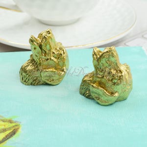 Può includere: Due figurine di animali in metallo dorato, forse cani, con la testa inclinata verso l'alto. Le figurine sono su una superficie turchese.