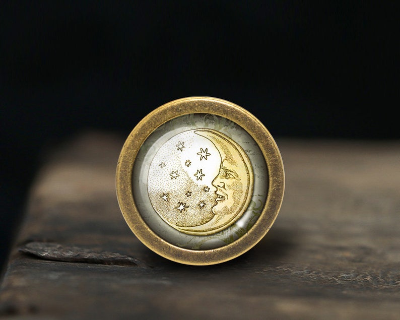 May include: A gold-toned metal ring with a glass cabochon featuring a crescent moon and stars design. The moon is depicted with a face and the stars are scattered around the moon.
