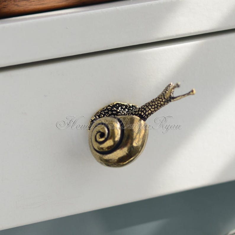 Puede incluir: Un tirador de caj&oacute;n en forma de caracol dorado con una concha detallada y una cola enroscada. El tirador est&aacute; montado en un frente de caj&oacute;n blanco.