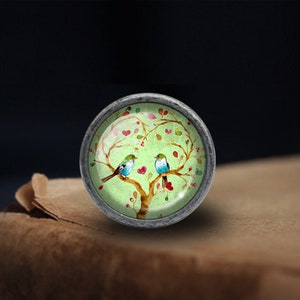 Puede incluir: Un pomo redondo plateado con un inserto de vidrio que presenta un &aacute;rbol verde con corazones rosas y dos p&aacute;jaros azules.