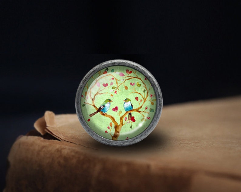 Puede incluir: Un pomo redondo plateado con un inserto de vidrio que presenta un &aacute;rbol verde con corazones rosas y dos p&aacute;jaros azules.
