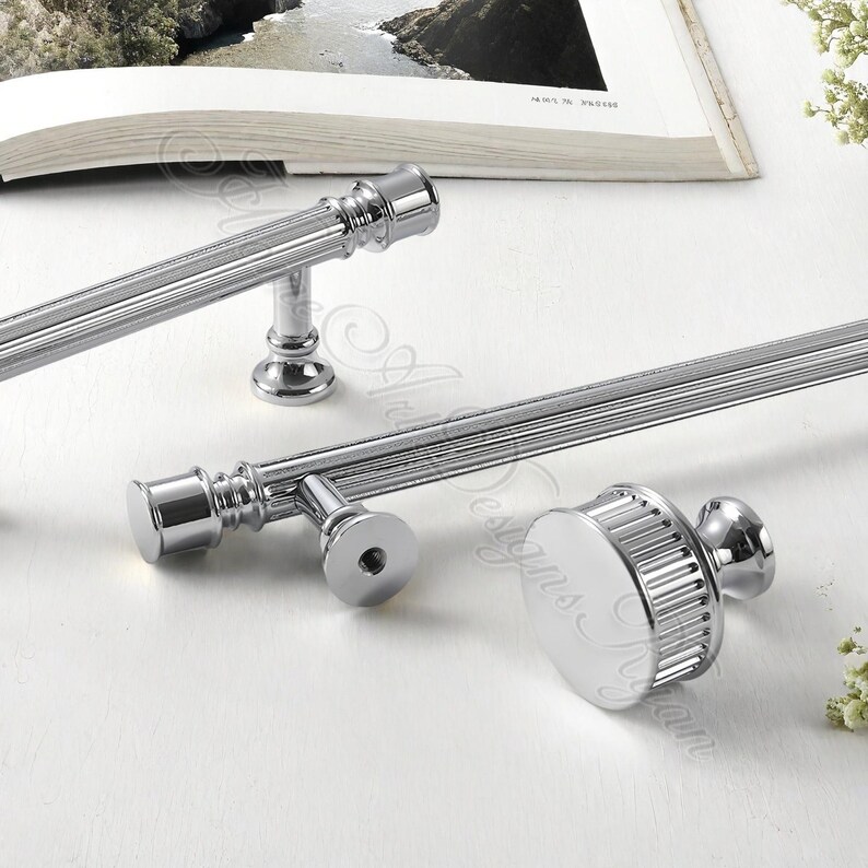 Puede incluir: Primer plano de varias piezas de herrajes de gabinete cromados pulidos. Los herrajes incluyen tiradores de barra largos y un pomo redondo, todos con un dise&ntilde;o acanalado. Las piezas est&aacute;n dispuestas sobre una superficie blanca, lo que sugiere un tema de decoraci&oacute;n del hogar.