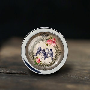 Peut inclure: Un bouton argenté avec un dessus rond en verre orné d'un motif vintage d'oiseaux et de fleurs. Le motif comprend une couronne et le mot "GRACE".