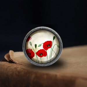 May include: A silver-toned round charm with a glass cabochon featuring a vintage-style illustration of red poppies on a cream background.