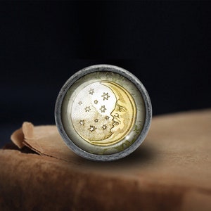 May include: A round, silver-toned knob with a glass insert featuring a gold crescent moon with a face and stars. The knob is on a brown surface.