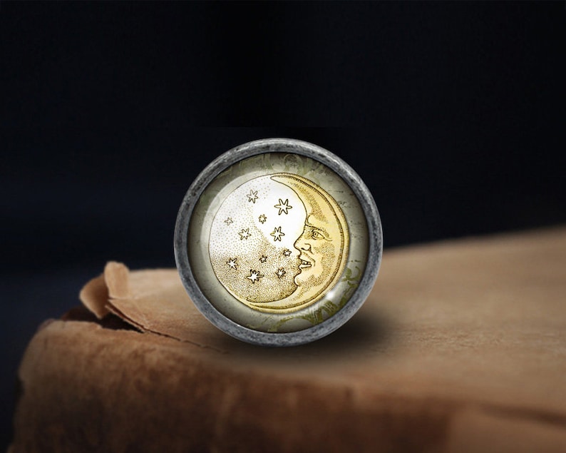 May include: A round, silver-toned knob with a glass insert featuring a gold crescent moon with a face and stars. The knob is on a brown surface.