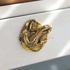 May include: Gold frog drawer pull with a detailed design. The frog is perched on a leaf with its legs outstretched.