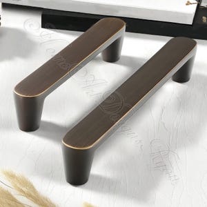 K&ouml;nnte beinhalten: Zwei bronzefarbene Schrankgriffe mit rechteckigem Design. Die Griffe haben eine dunkelbraune Oberfl&auml;che mit goldfarbenem Rand an den Kanten. Die Griffe sind auf einer wei&szlig;en Oberfl&auml;che montiert.