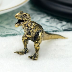 May include: A small, gold-coloured, metal Tyrannosaurus Rex dinosaur figurine. The figurine is standing on a white surface.