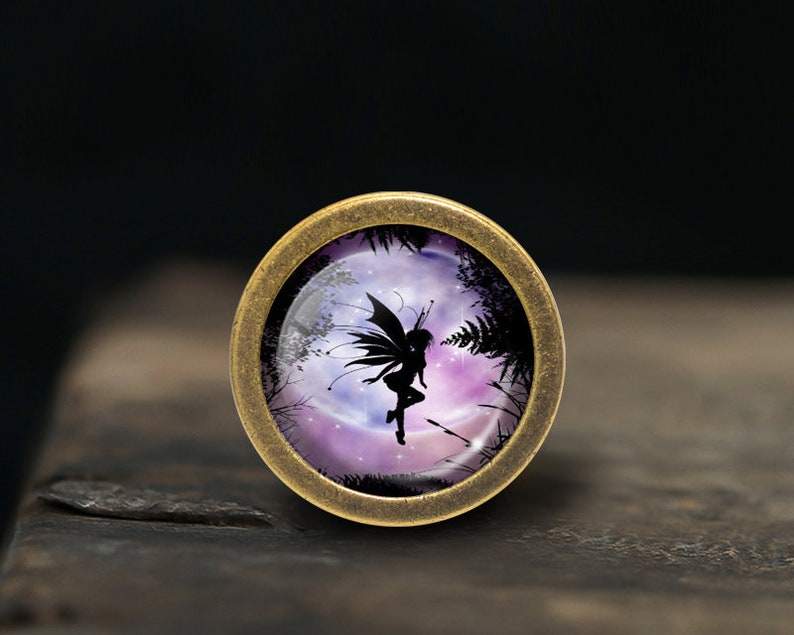 May include: A round, bronze-colored pendant with a glass insert featuring a silhouette of a fairy flying in front of a full moon. The moon is surrounded by a purple and pink nebula with stars.