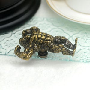 Pu&ograve; includere: Un pomello per armadio in bronzo, dettagliato e scolpito a forma di creatura scimmiesca muscolosa e pelosa. La creatura &egrave; posizionata sulla schiena con le braccia alzate sopra la testa.