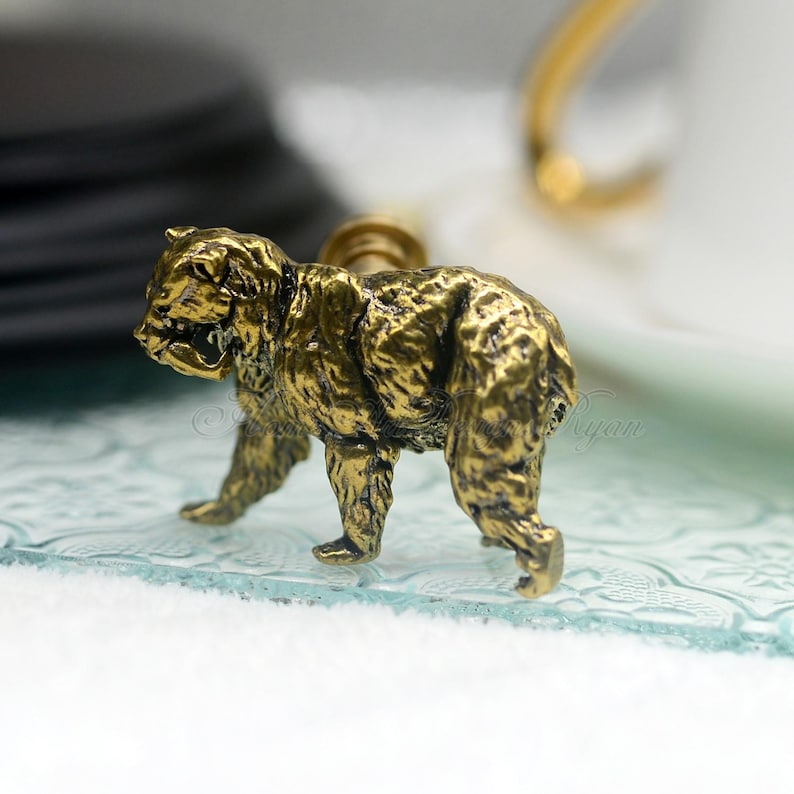 May include: A gold-colored bear figurine with a detailed textured surface. The bear is standing on a glass surface with a decorative pattern.