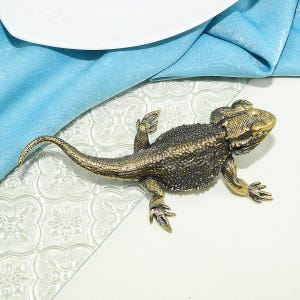 Può includere: Una statuetta in metallo dorato raffigurante un drago barbuto. Il drago è in posa con la coda arrotolata e la testa inclinata di lato. La statuetta è su una superficie bianca con uno sfondo a motivi blu e bianchi.