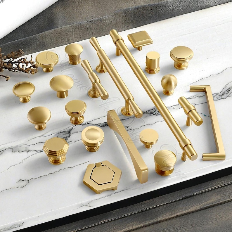 Puede incluir: Una colecci&oacute;n de herrajes dorados para gabinetes, que incluye perillas y tiradores, dispuestos sobre una superficie con dise&ntilde;o de m&aacute;rmol. Los herrajes presentan varias formas, como estilos redondos, cuadrados y de barra, ofreciendo opciones de dise&ntilde;o.