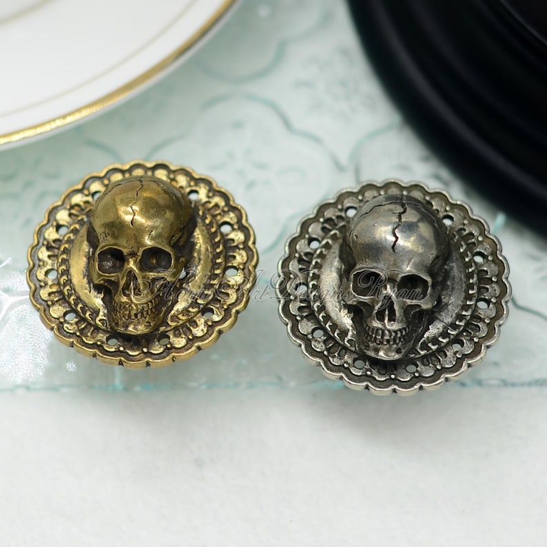 May include: Two decorative skull-shaped cabinet knobs. One is gold-toned, the other silver-toned. Each skull is centered on a round, ornate, filigree-style base. The knobs are likely metal and designed for furniture. The knobs are suitable for furniture.