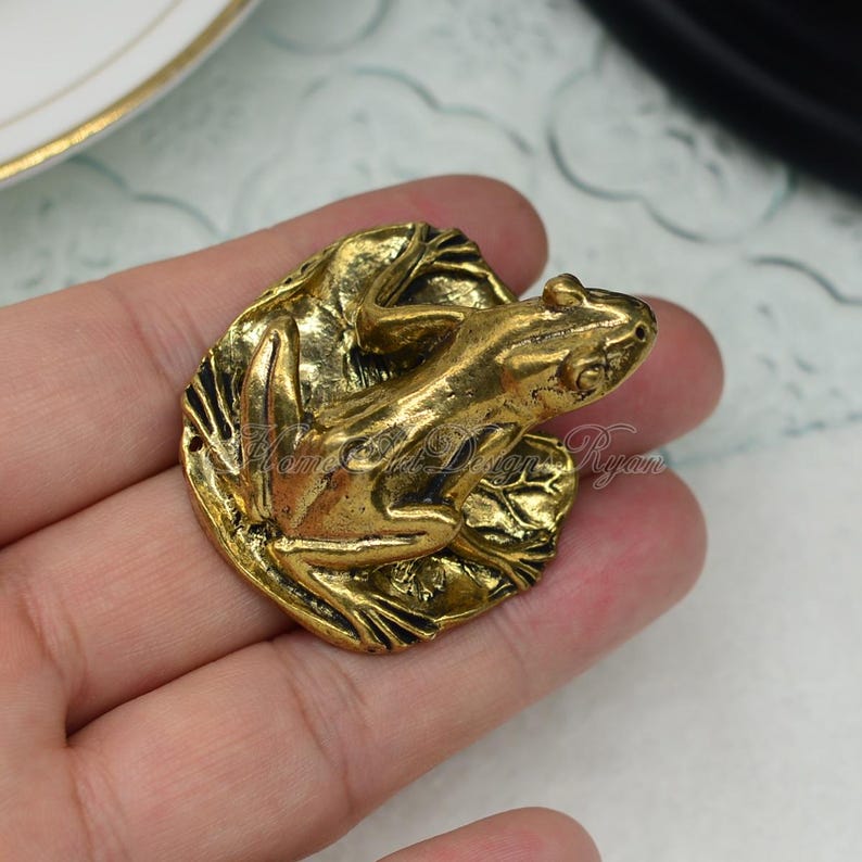 May include: A gold-toned frog figurine sitting on a leaf. The frog is detailed with textured skin and a realistic pose.