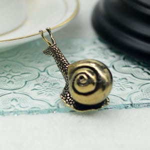 Puede incluir: Una figurita de caracol de metal dorado con una concha detallada y un cuerpo texturizado. El caracol est&aacute; sentado sobre una superficie de vidrio azul claro.